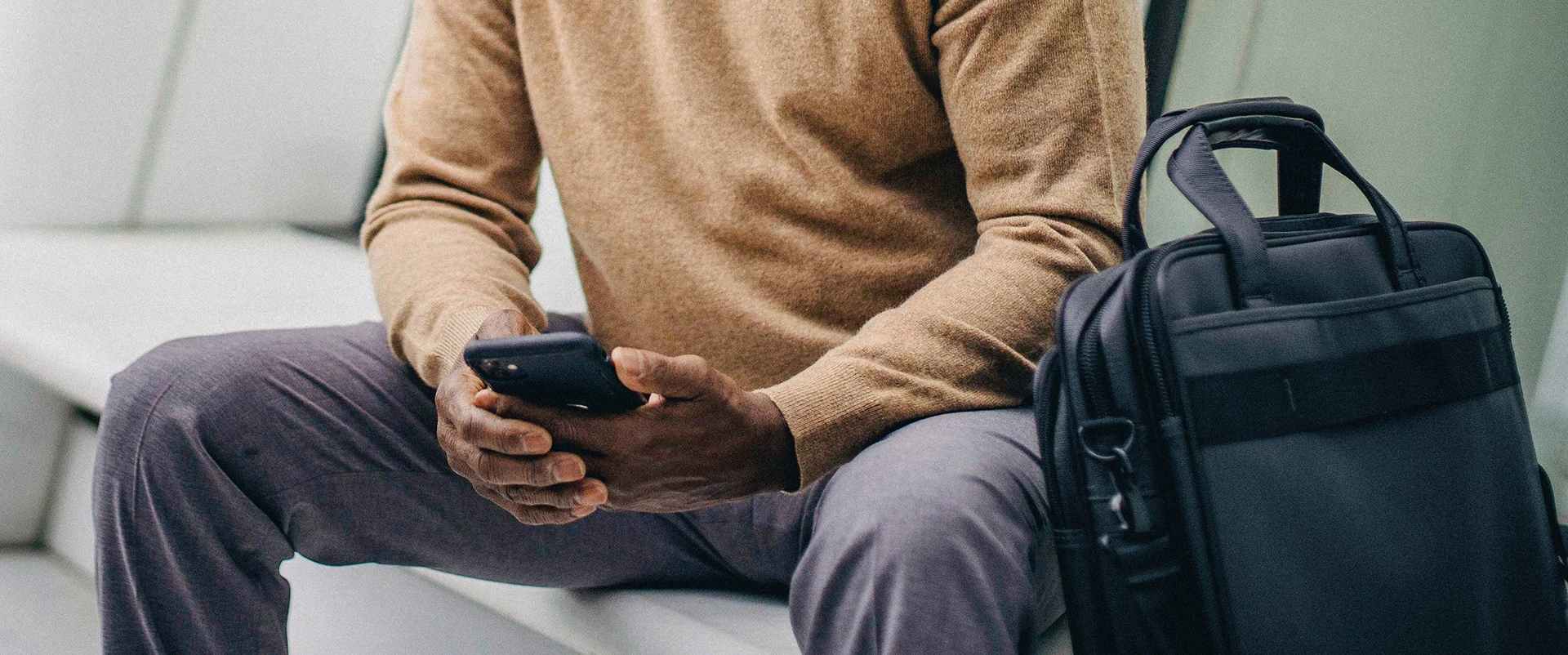 Program evaluation professional reviewing data on smartphone while sitting on couch next to black leather laptop bag, wearing beige sweater